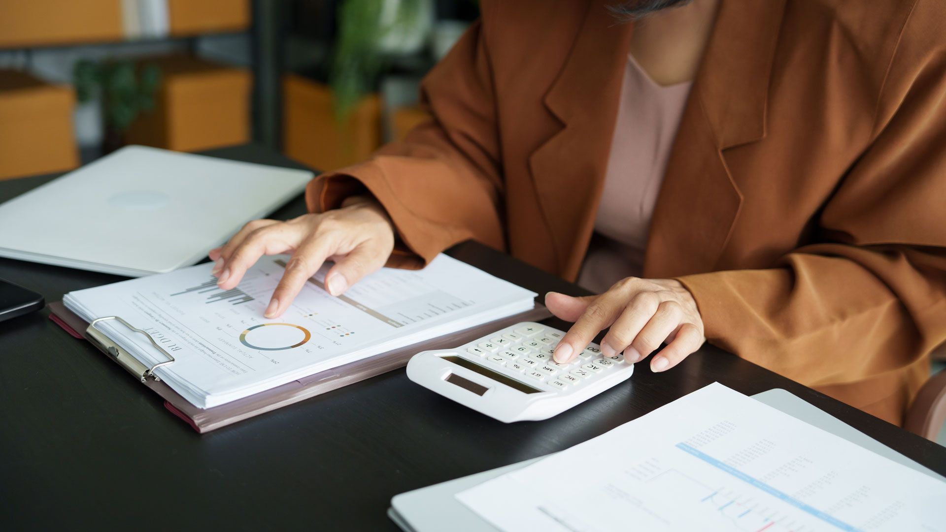 Person in brown blazer using calculator and pointing at a graph on a document.
