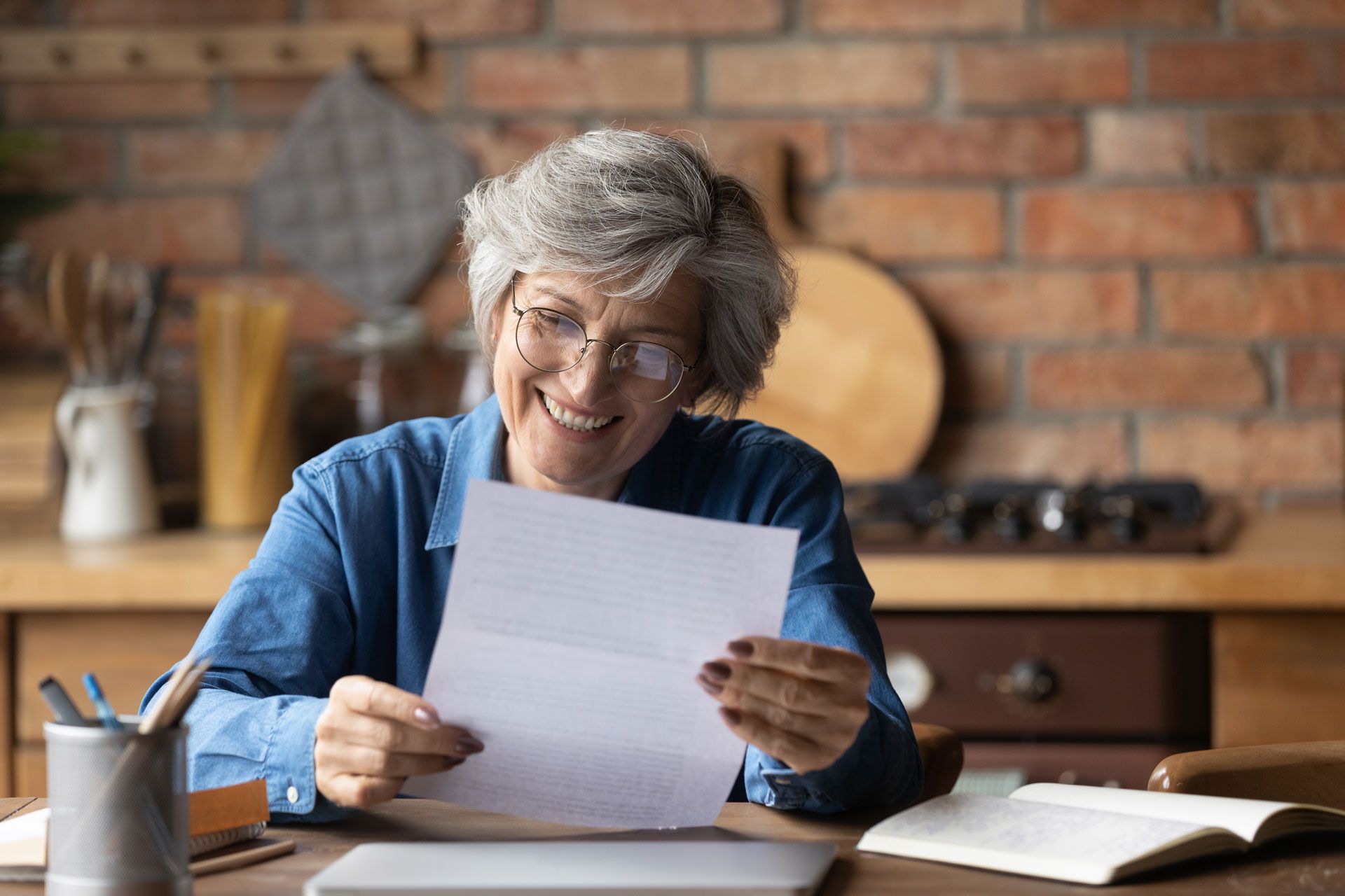 Woman with glasses smiles while reading a letter at a table with a laptop, pens, and a notebook in a kitchen.