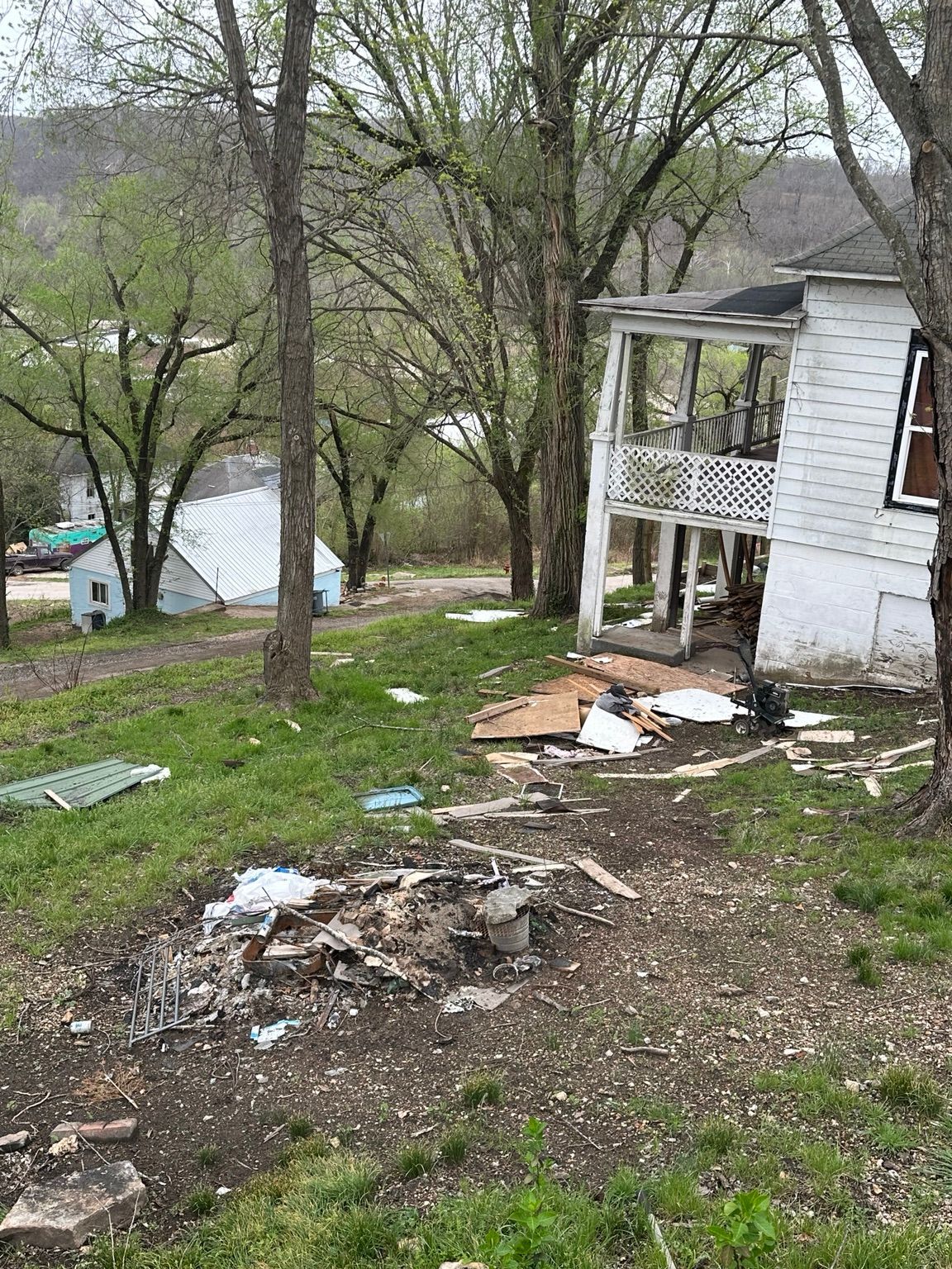 Damaged white house porch on a hillside with debris and a charred fire pit in the yard