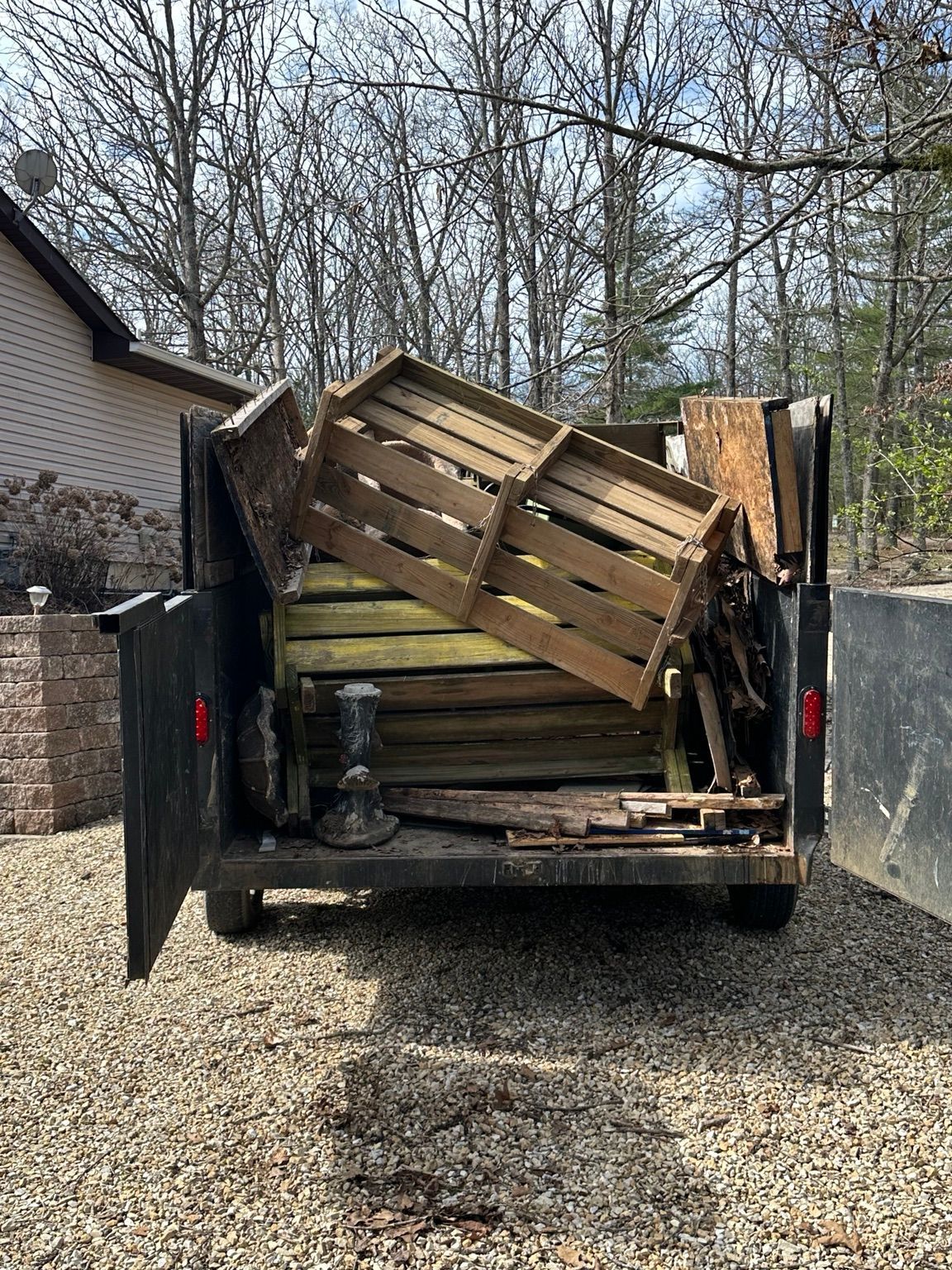 Trailer loaded with stacked wooden pallets and boards, rear doors open on a gravel driveway.