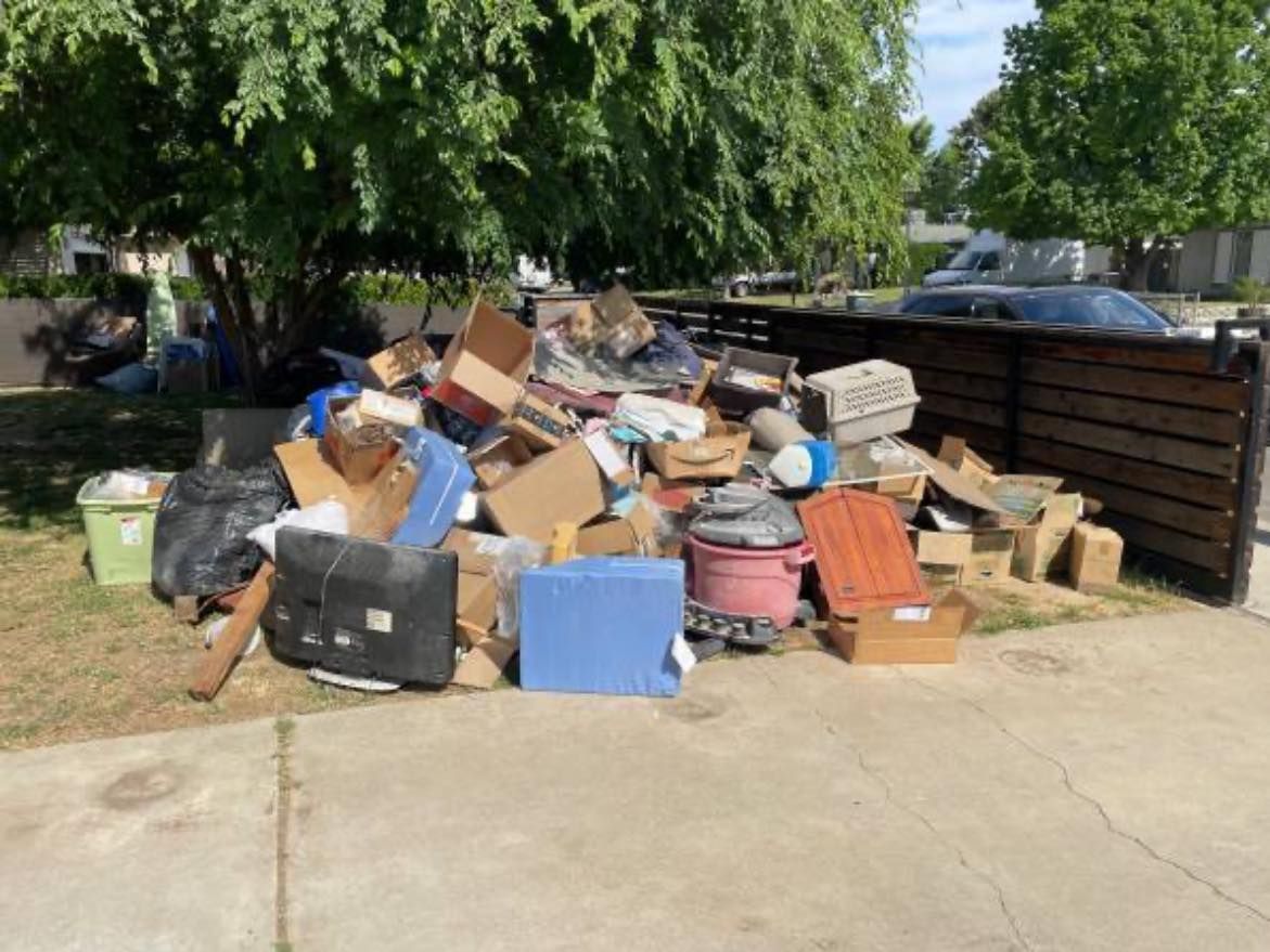 Overflowing pile of mixed trash and discarded furniture on a sidewalk beside a wooden fence and trees