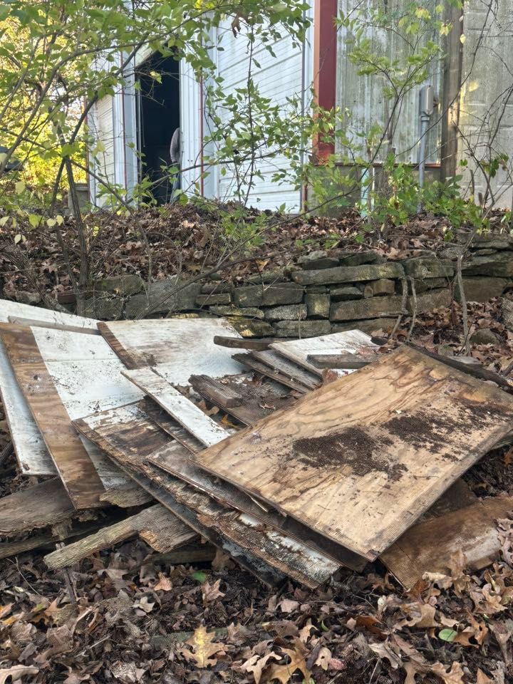 Piles of broken wood boards and debris stacked in front of a stone wall outdoors under trees