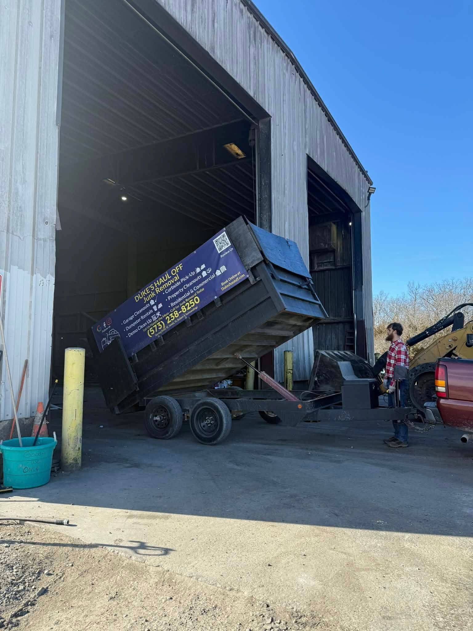 Dump trailer raised at a warehouse entrance, with workers nearby and a red vehicle beside it.