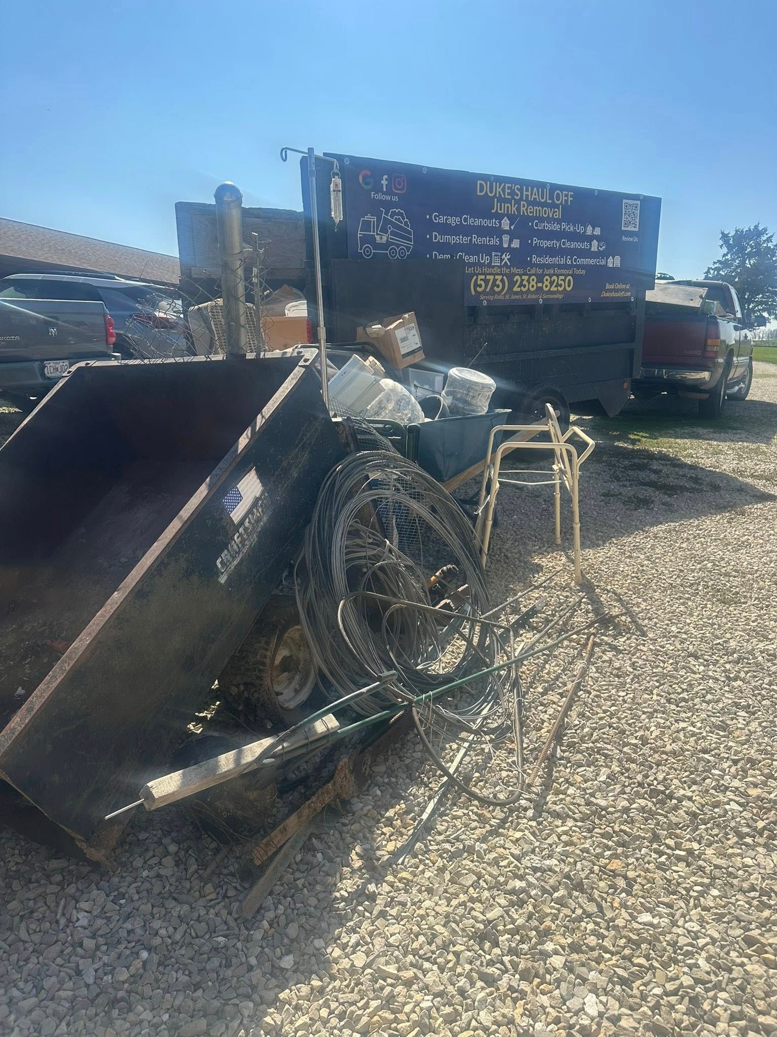 Damaged black trailer with bent wheel in a gravel lot beside a large truck under a blue sky