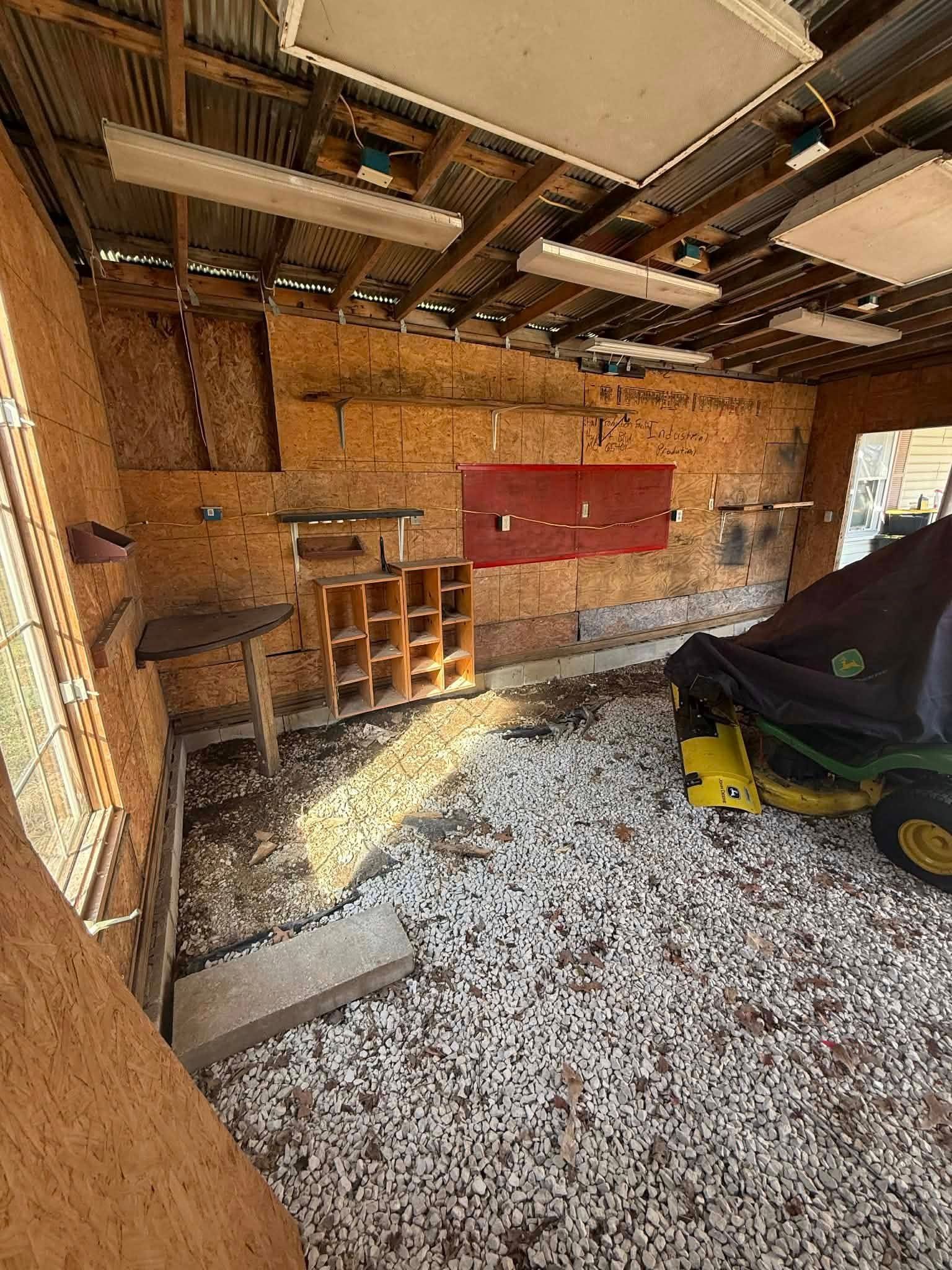 Garage with exposed insulation, red wall cabinets, shelves, and a parked riding mower on gravel.