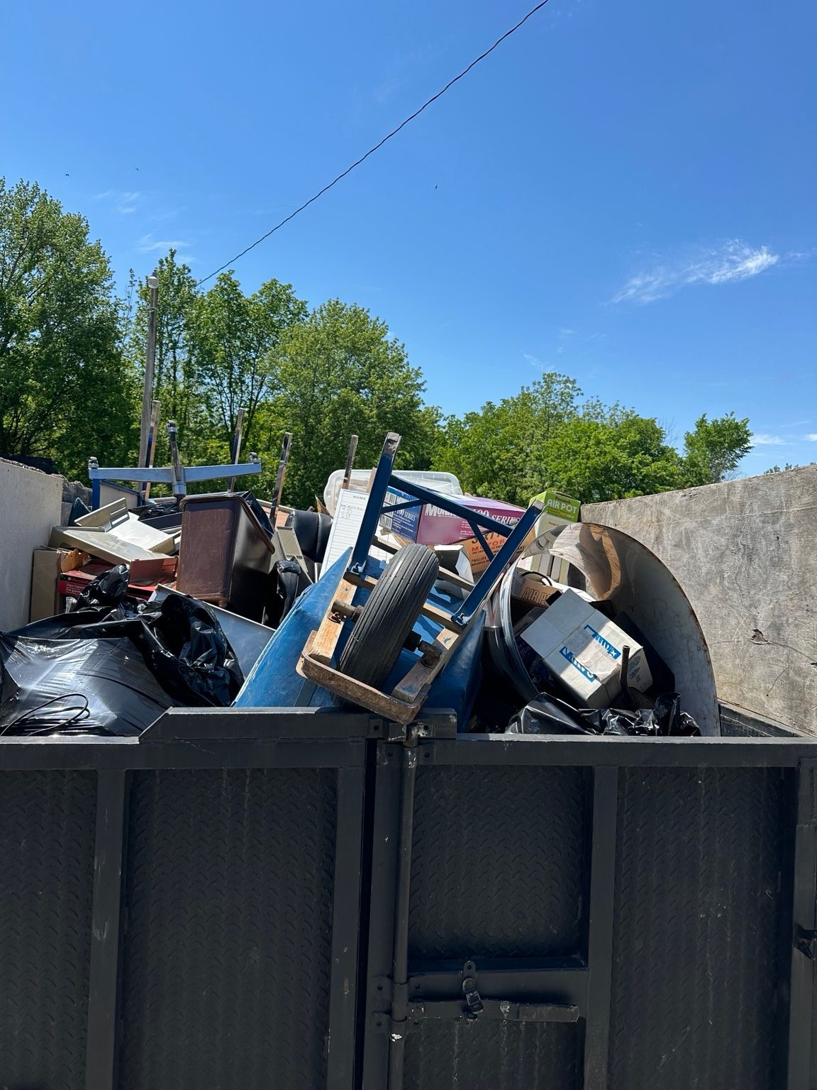 Dumpster filled with scrap metal, trash bags, and debris outdoors under a blue sky