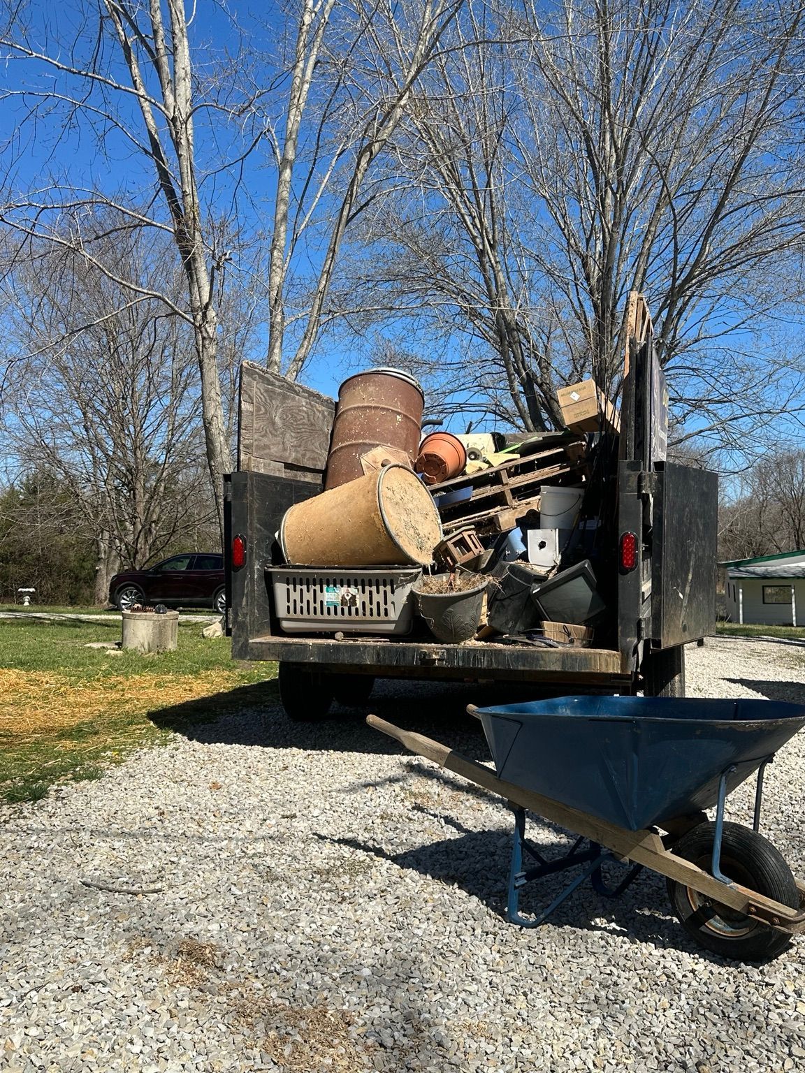Pickup truck loaded with discarded furniture and debris beside a wheelbarrow on a gravel driveway