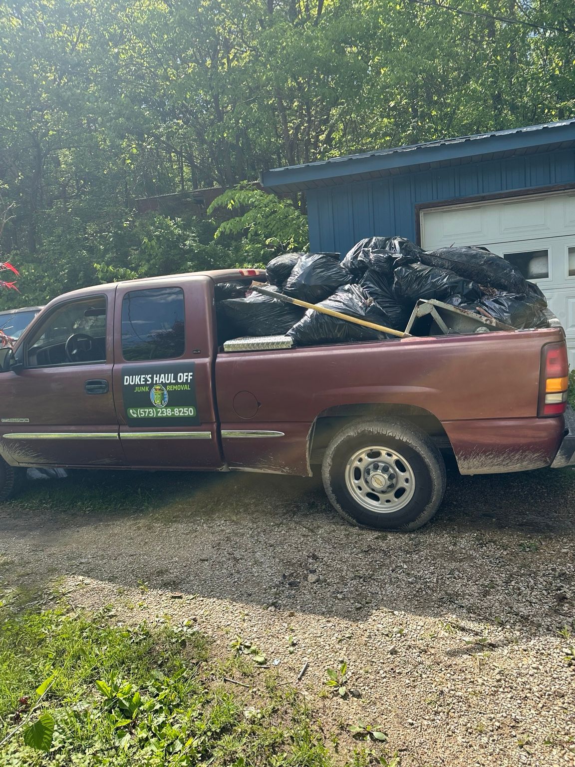 Maroon pickup truck loaded with black debris parked beside a blue building on a gravel lot