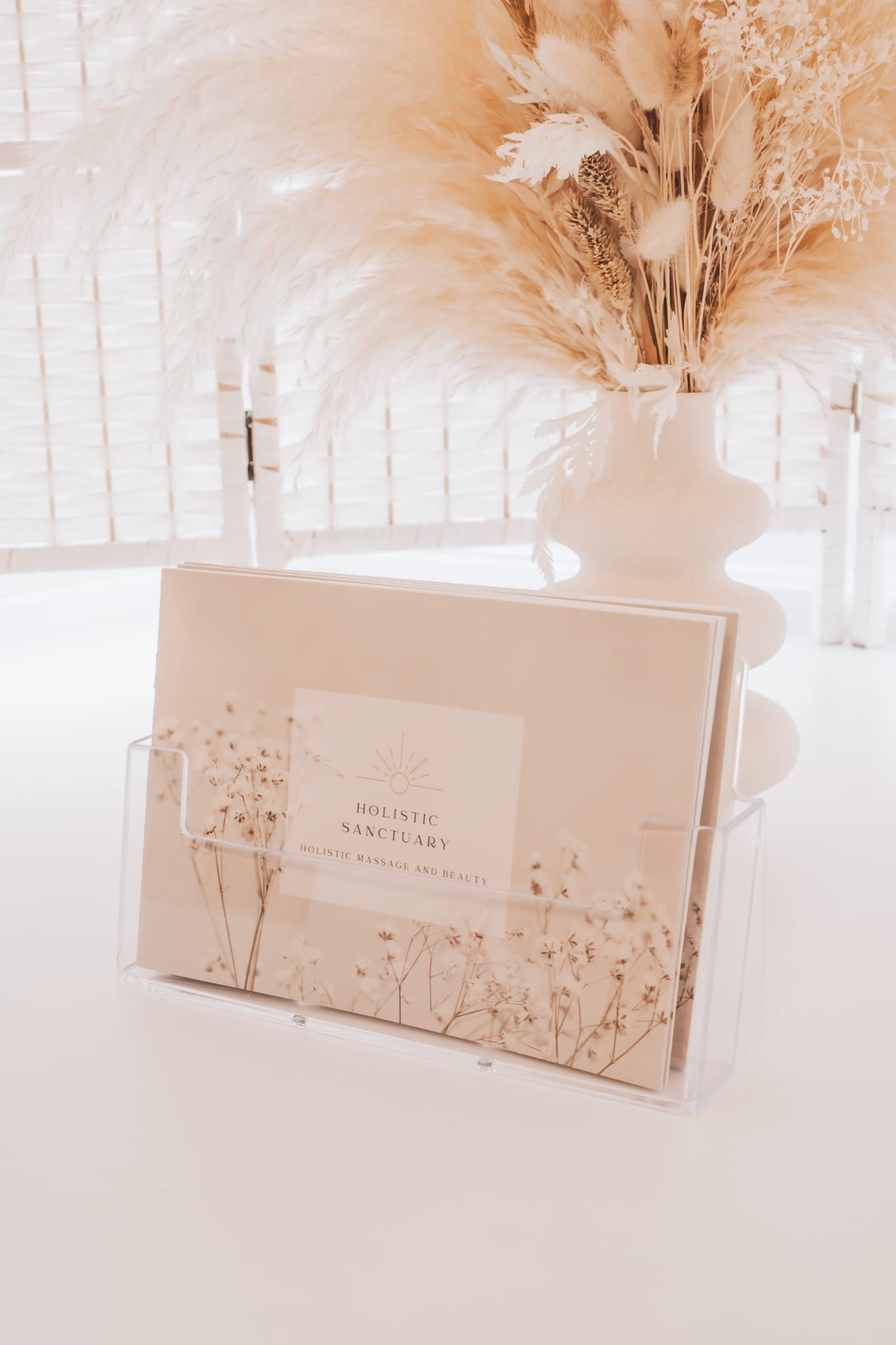 Clear acrylic wall file holding stationery on a white surface, with beige dried flowers in a white vase.