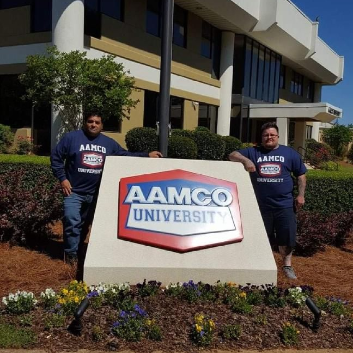 Two people stand beside an AAMCO University sign outside a building with landscaping.