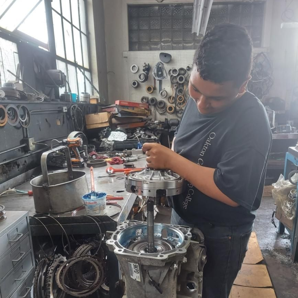 Mechanic working on an engine in a cluttered workshop, tools and metal parts around them.