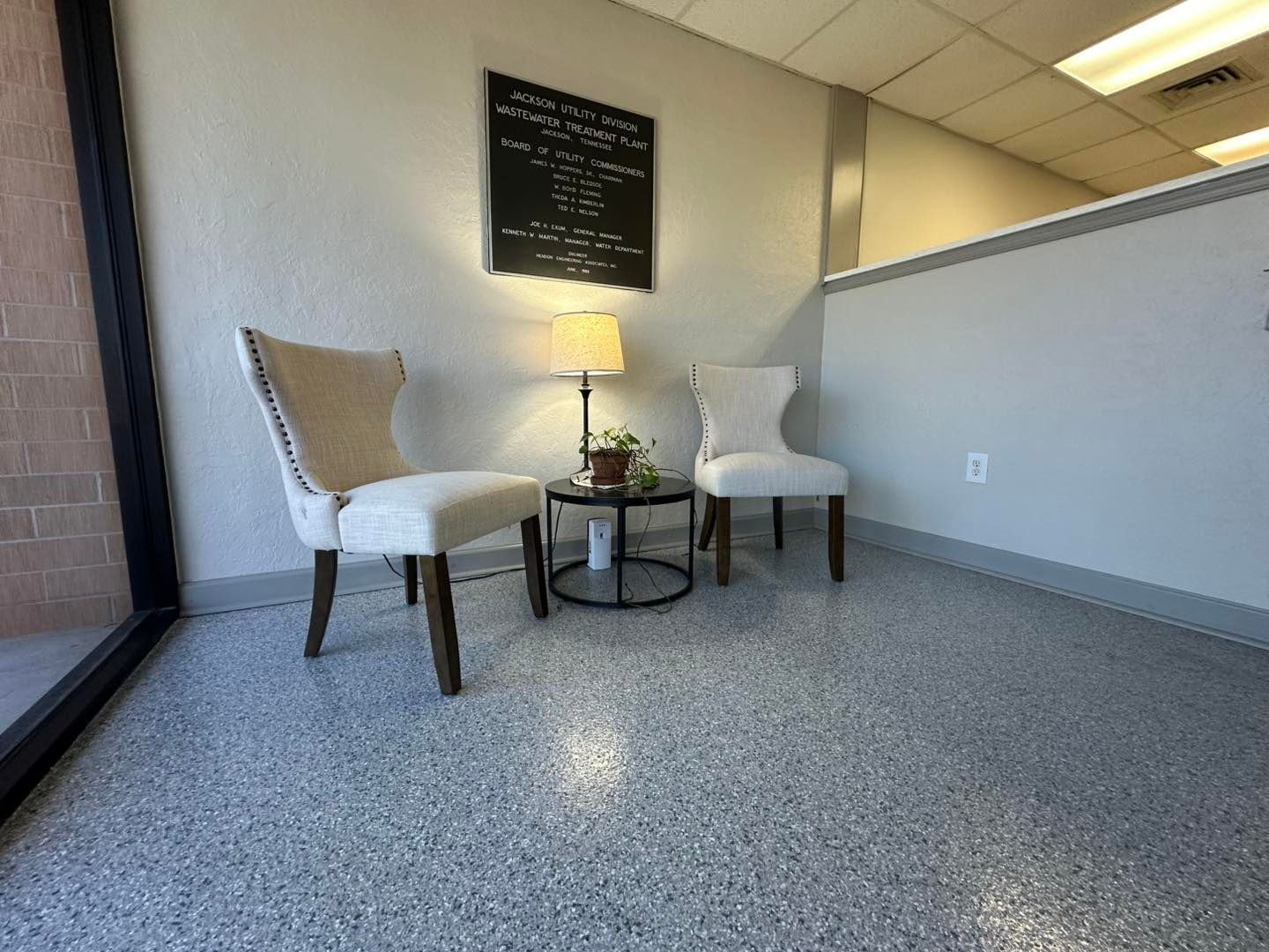 Waiting area with two chairs, small table, lamp, and framed artwork on speckled floor.