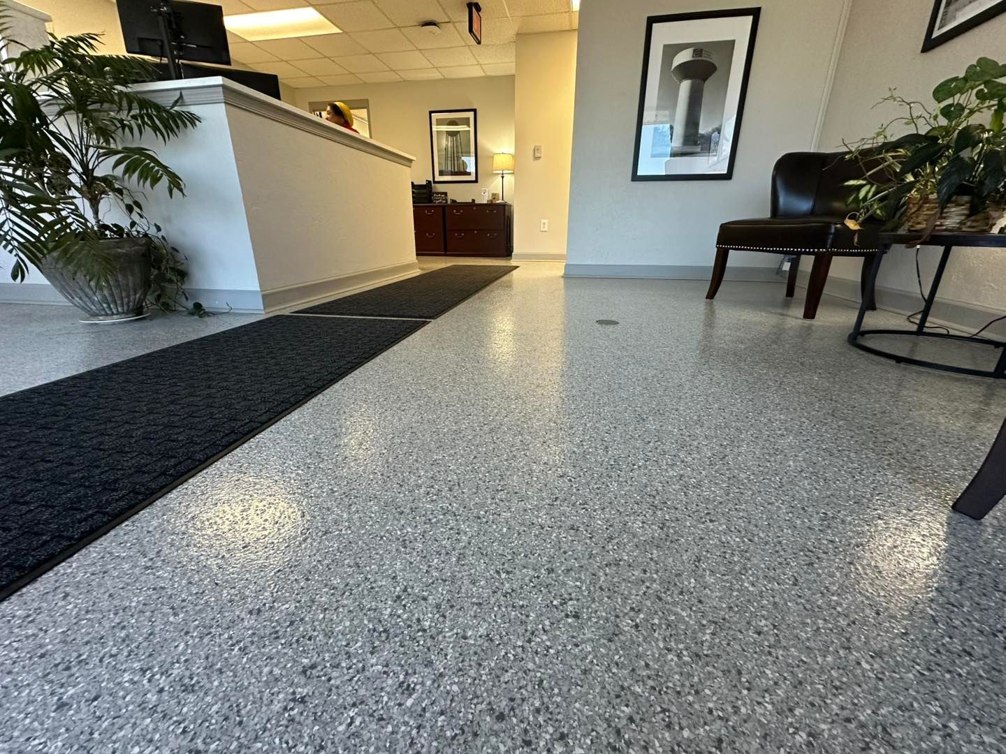 Office lobby with speckled gray floor and black entrance mat. Plants and seating are present.