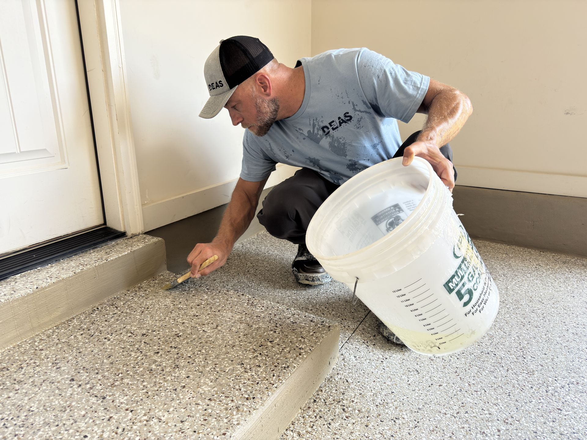 Man applying coating to a speckled garage floor, holding a bucket and trowel near a doorway.