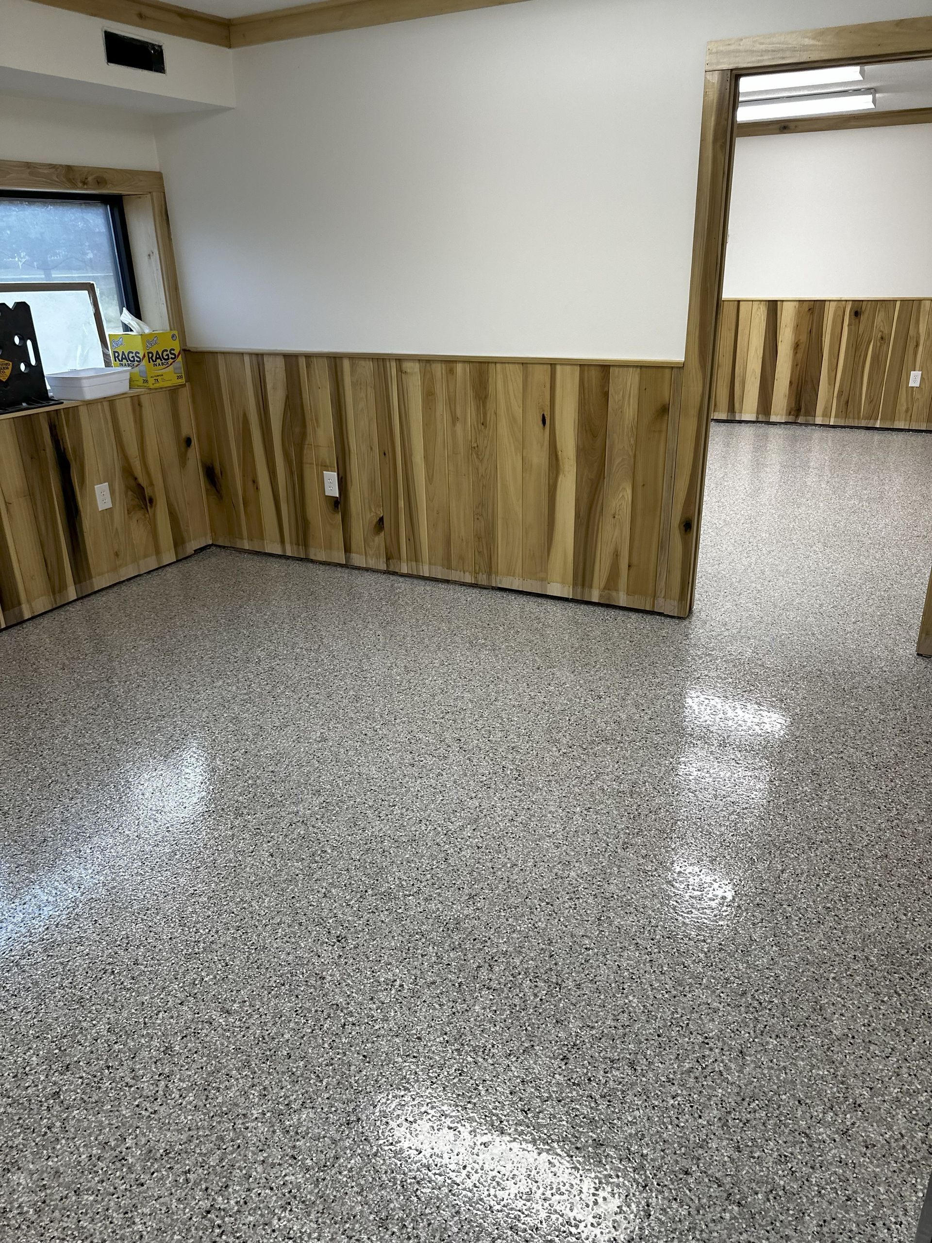 Room with wood paneling on the walls and speckled epoxy flooring. Another room visible through a doorway.