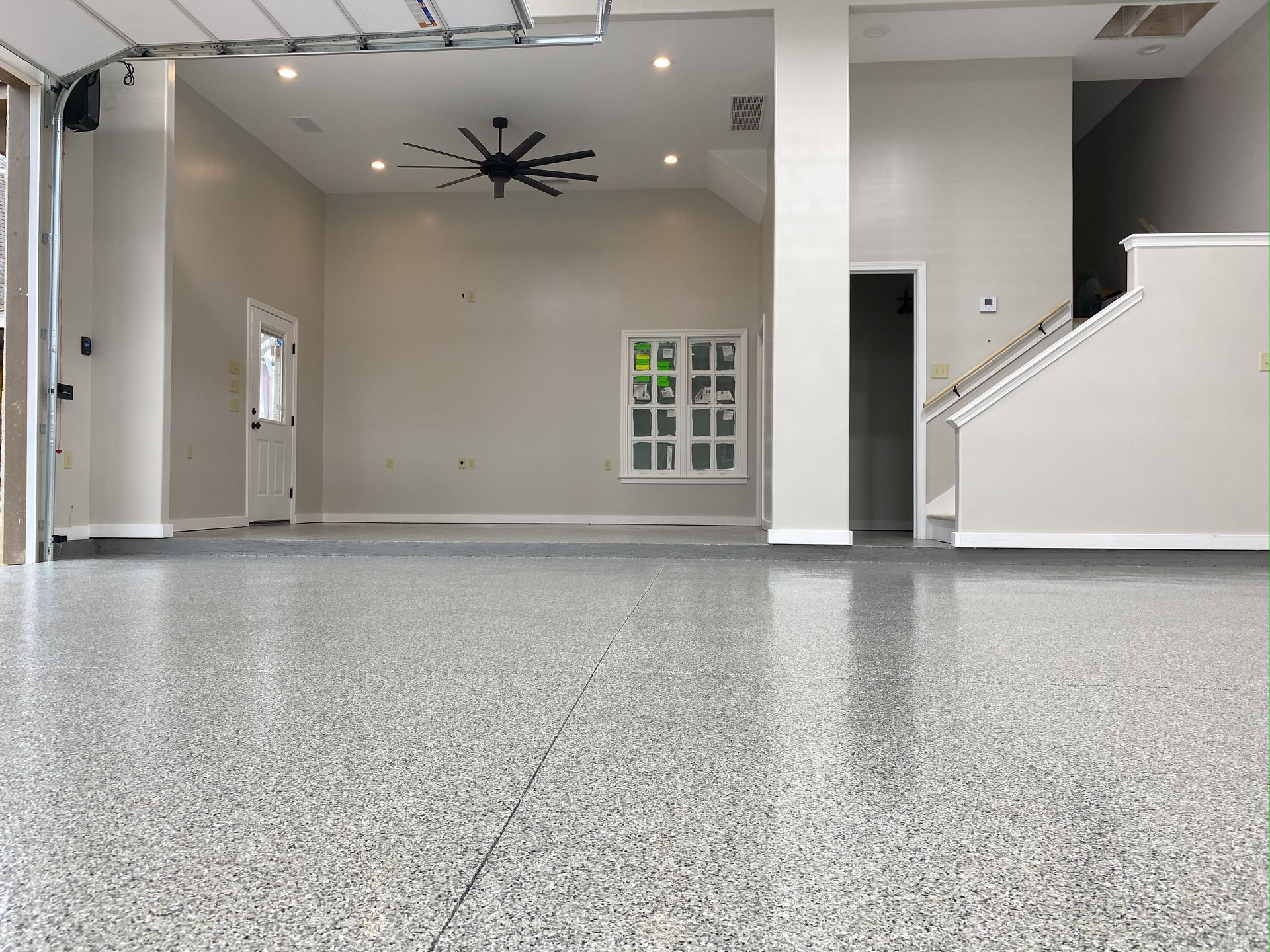 Gray epoxy-coated garage floor with an open door, a ceiling fan, and access to a staircase.