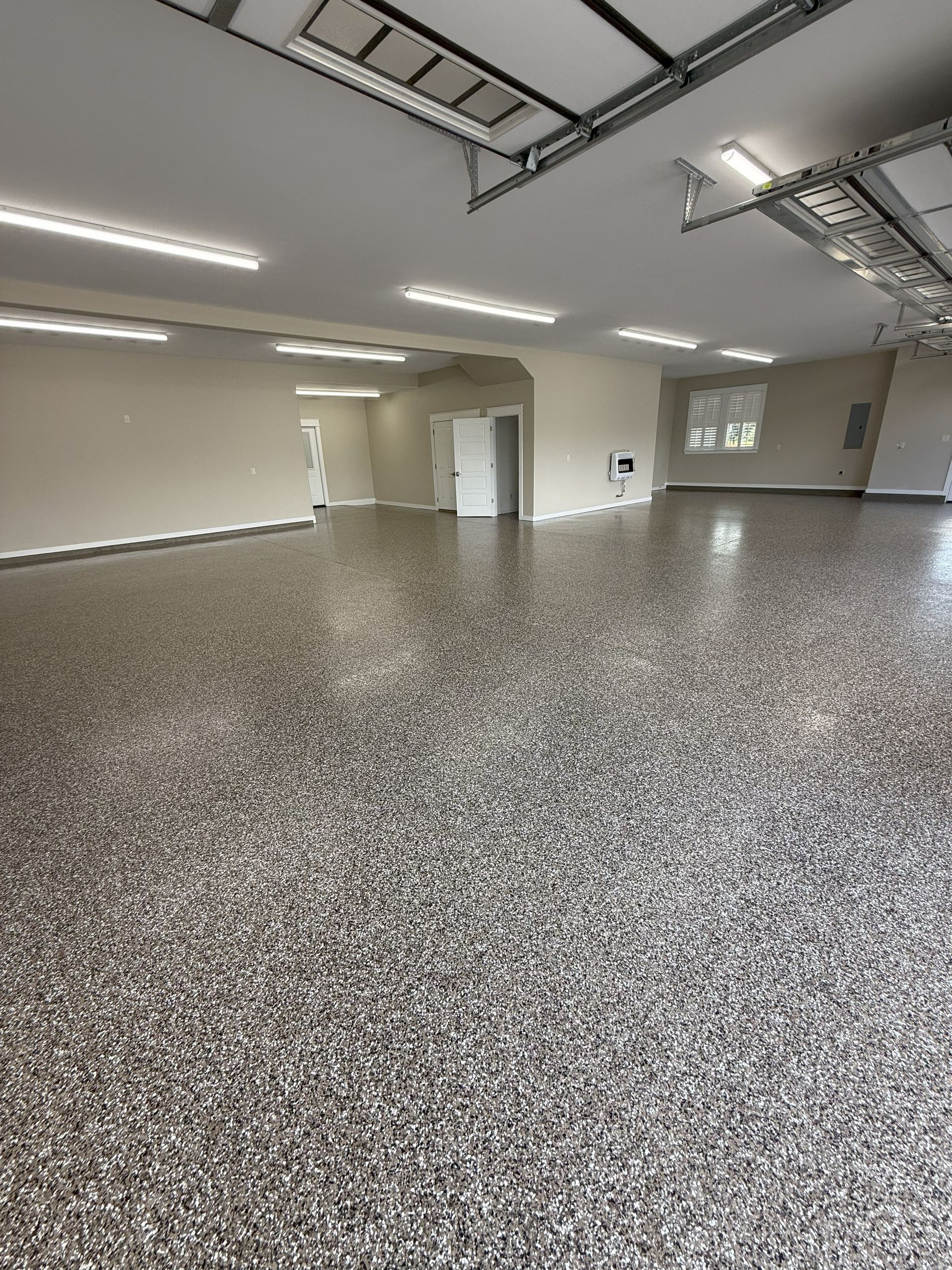 Large, empty garage with epoxy flooring and overhead lighting. Beige walls, white door, and garage door visible.