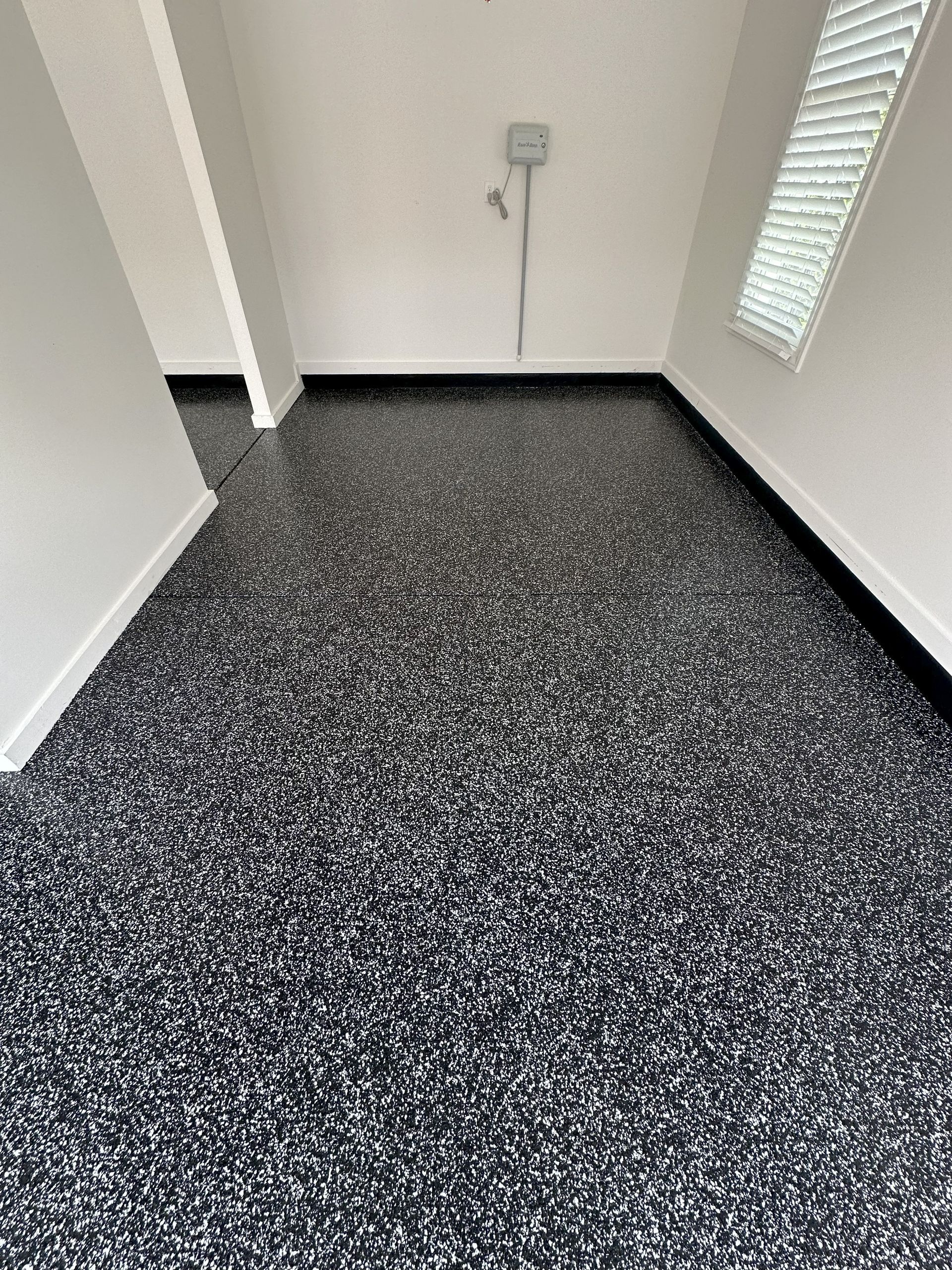 Garage interior with speckled black and white floor coating. White walls, black baseboards, and a window.