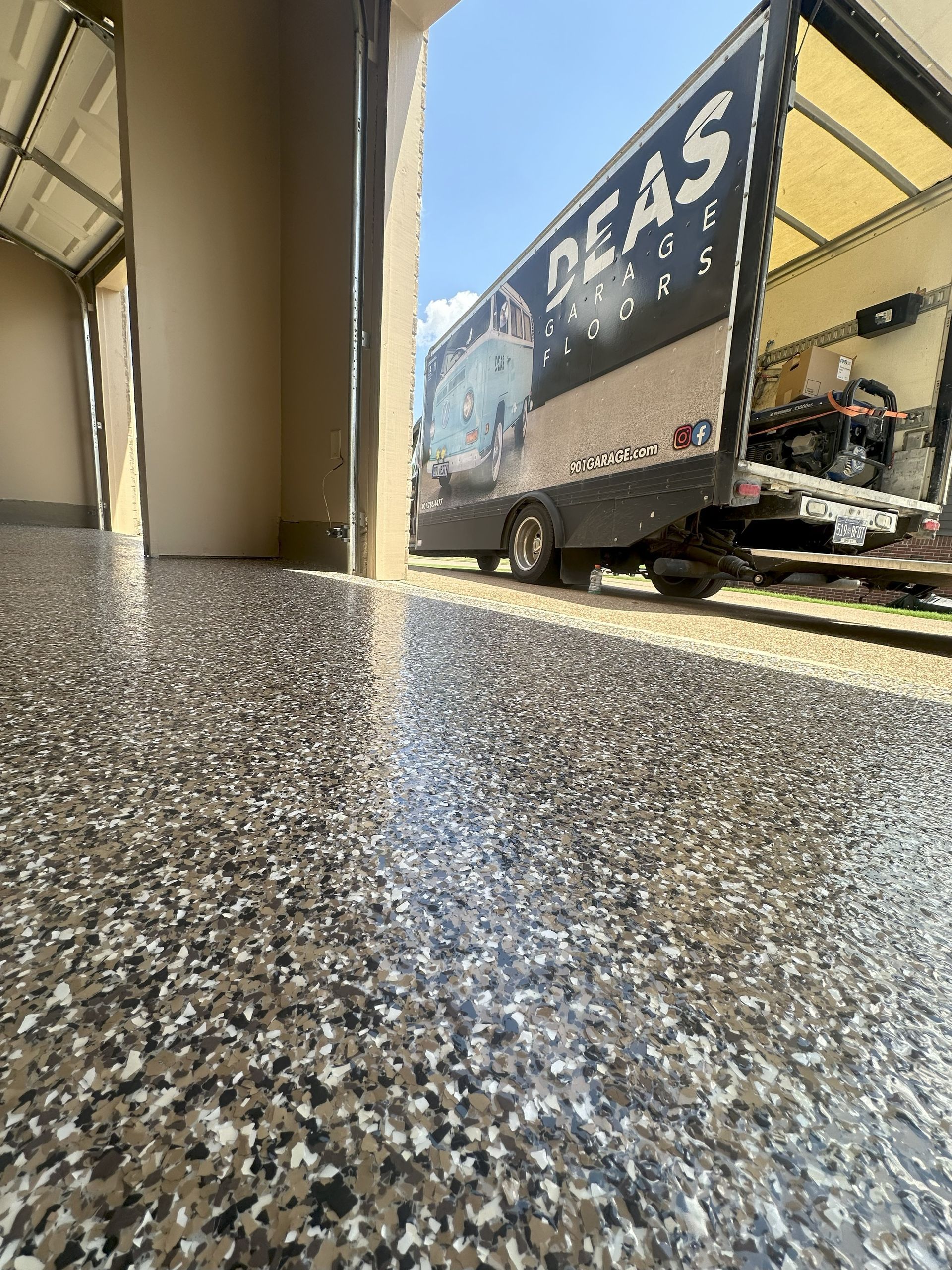 Garage floor with speckled epoxy finish. A trailer is parked outside. Sunny day.