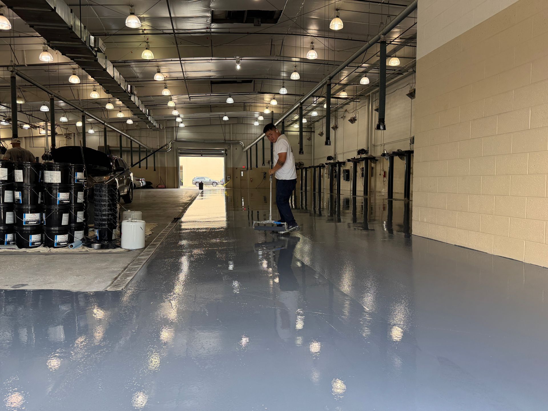 Person sweeping shiny, gray warehouse floor with rows of equipment on the wall, bright lights overhead.