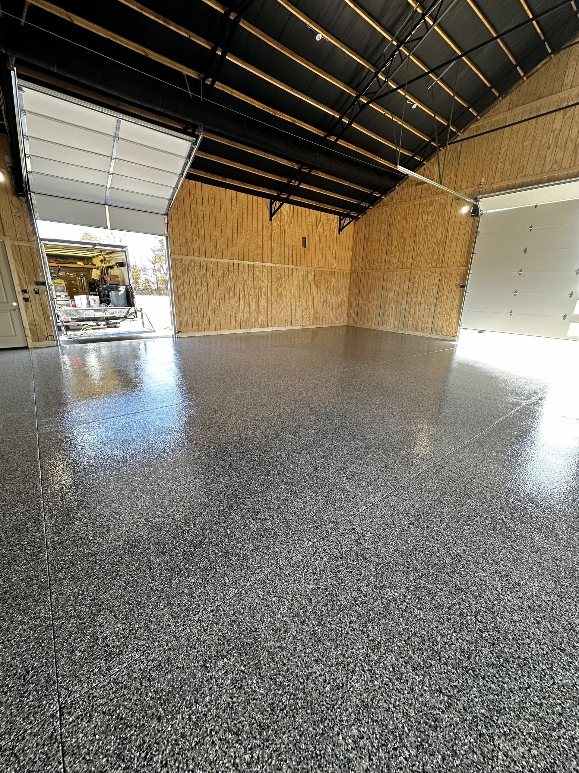 Garage with gray and black speckled epoxy floor, open door, wood walls, and black ceiling.