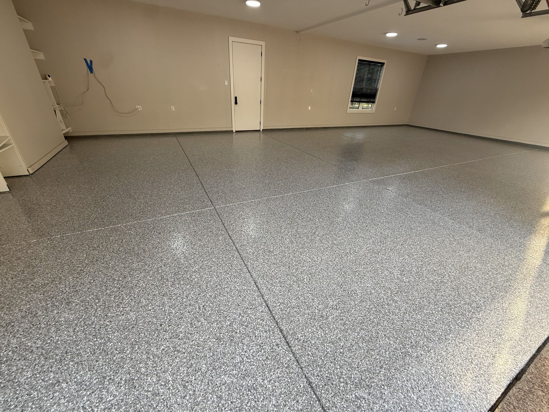 Newly coated garage floor with gray speckled epoxy. Beige walls, white door, and recessed lights.