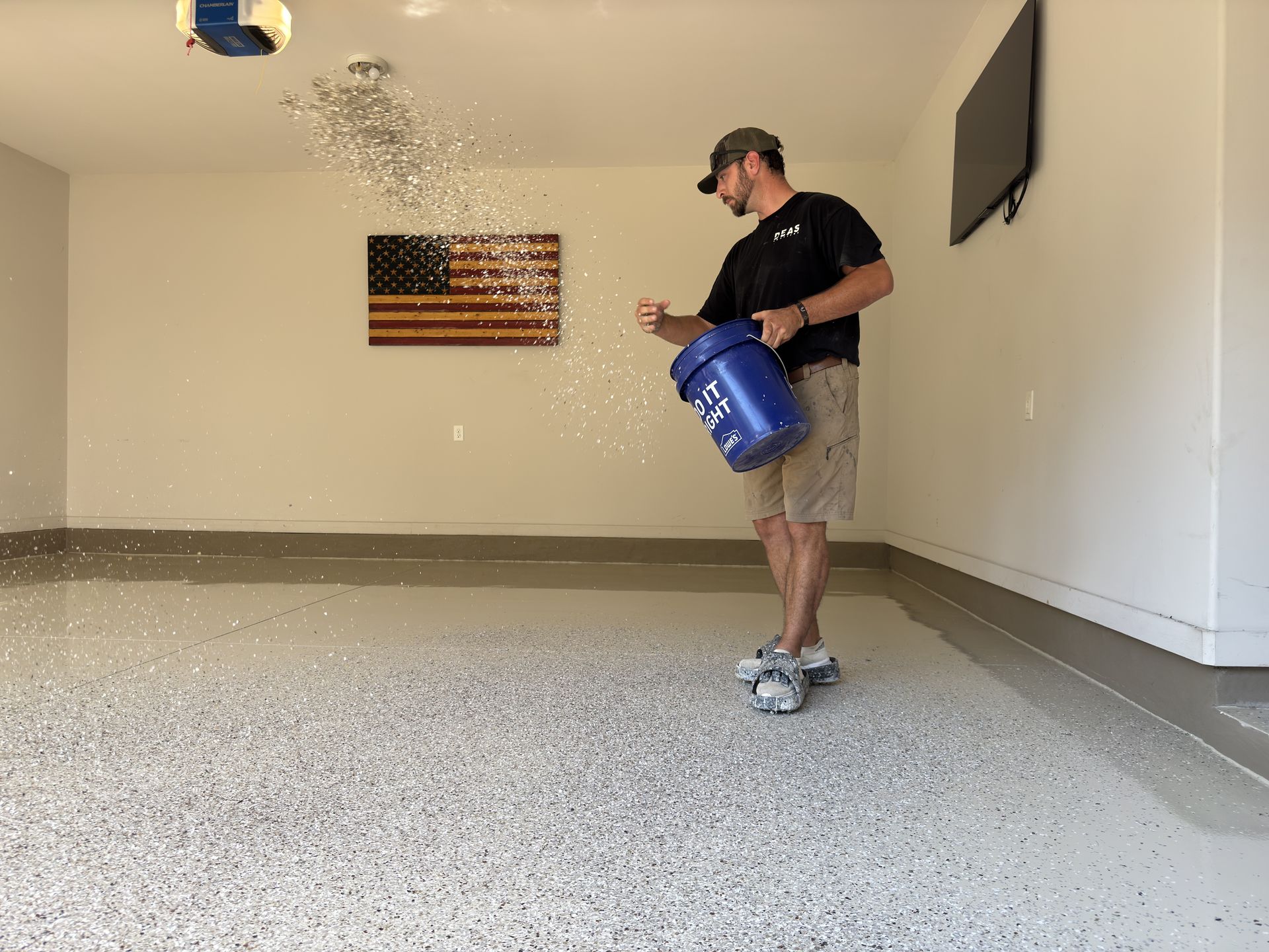 Man in garage sprinkles floor flakes from a blue bucket onto a partially covered epoxy floor.