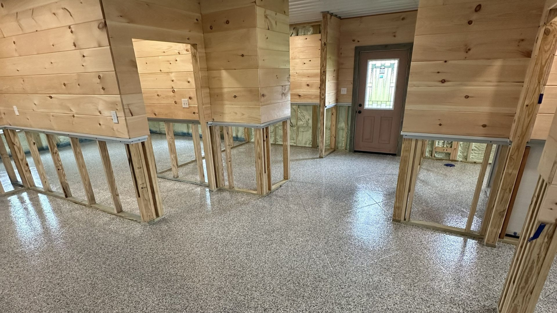 Interior construction site with wooden framing, paneling, and a speckled floor. Door and exposed insulation are visible.