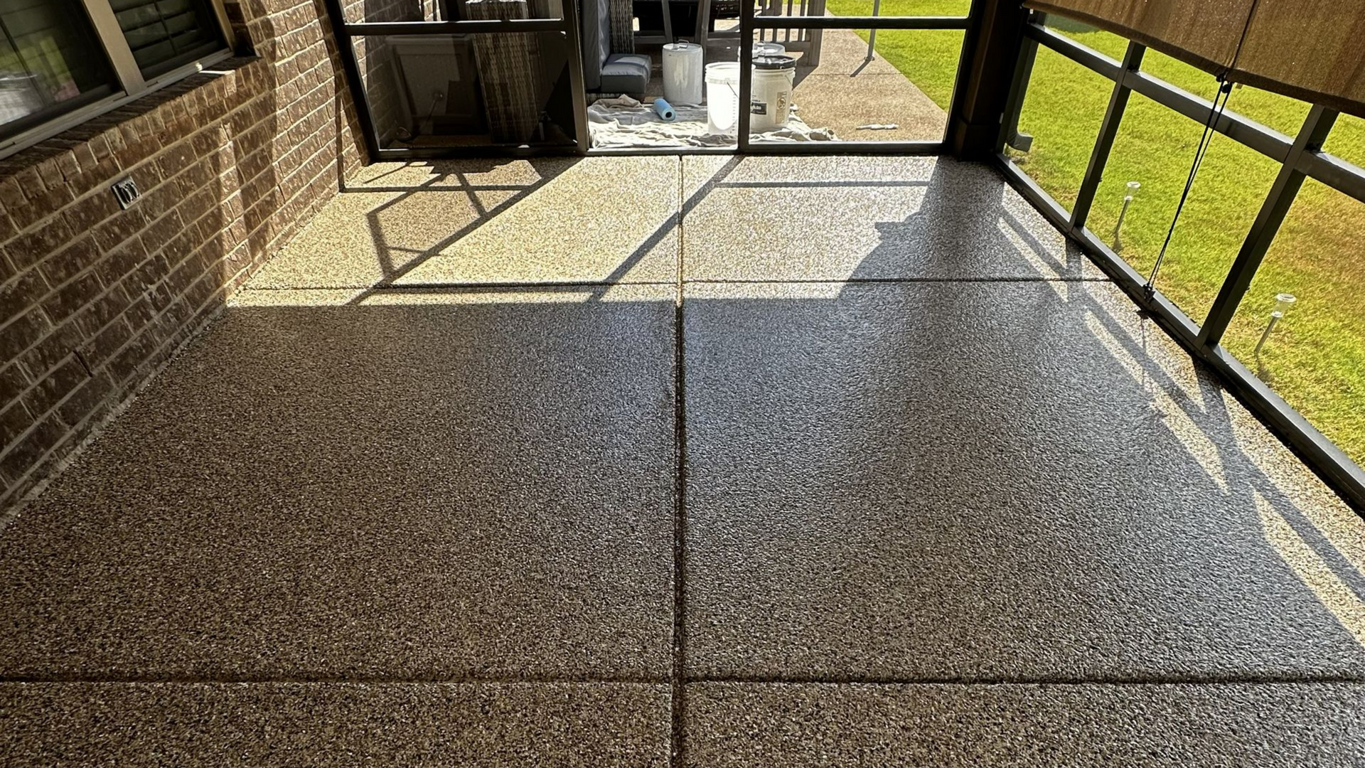 Sunlit covered patio with speckled concrete floor, black railings, and a gate opening to a grassy yard.