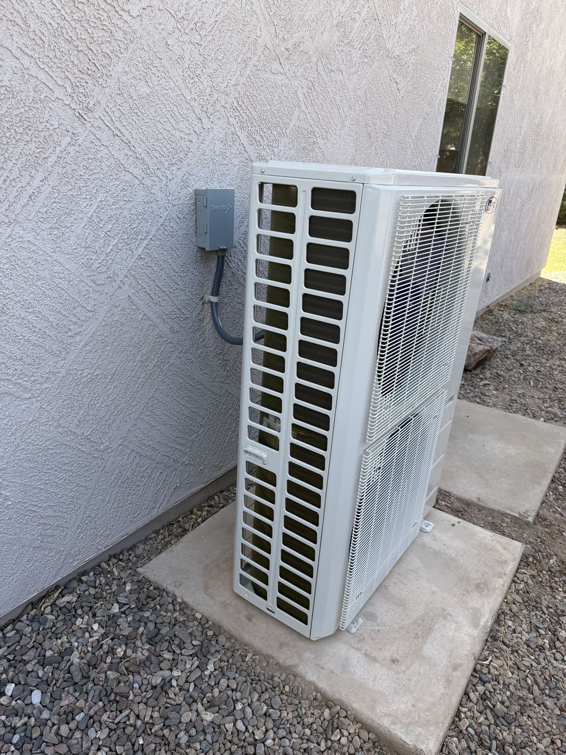 Outdoor air conditioner unit beside a stucco wall on a concrete pad with gravel nearby