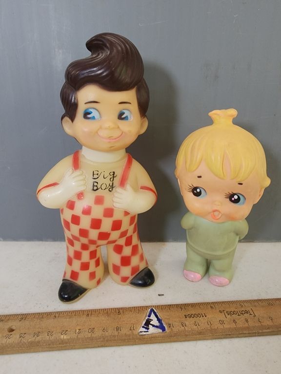 Two small cartoon figurines on a tabletop beside a ruler, one in red-checkered overalls and one in green.