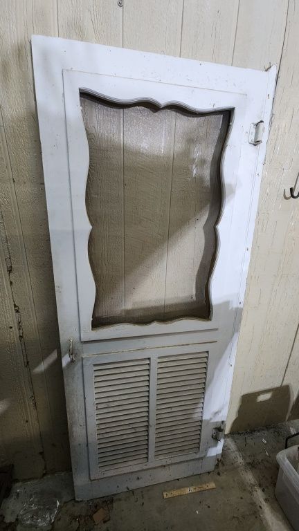 Damaged white door with a shattered upper window and vented lower panel leaning against a wall