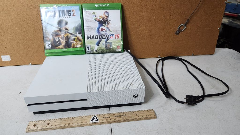 White Xbox One console with two game cases, cable, and ruler on a corkboard-backed surface
