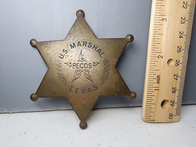 Brass U.S. Marshal star badge beside a wooden ruler on a white surface