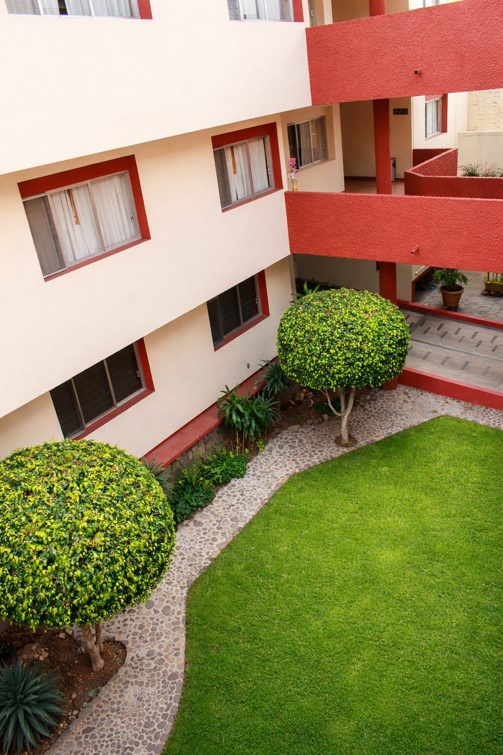 Patio con césped verde, árboles podados y edificio con detalles en rojo.