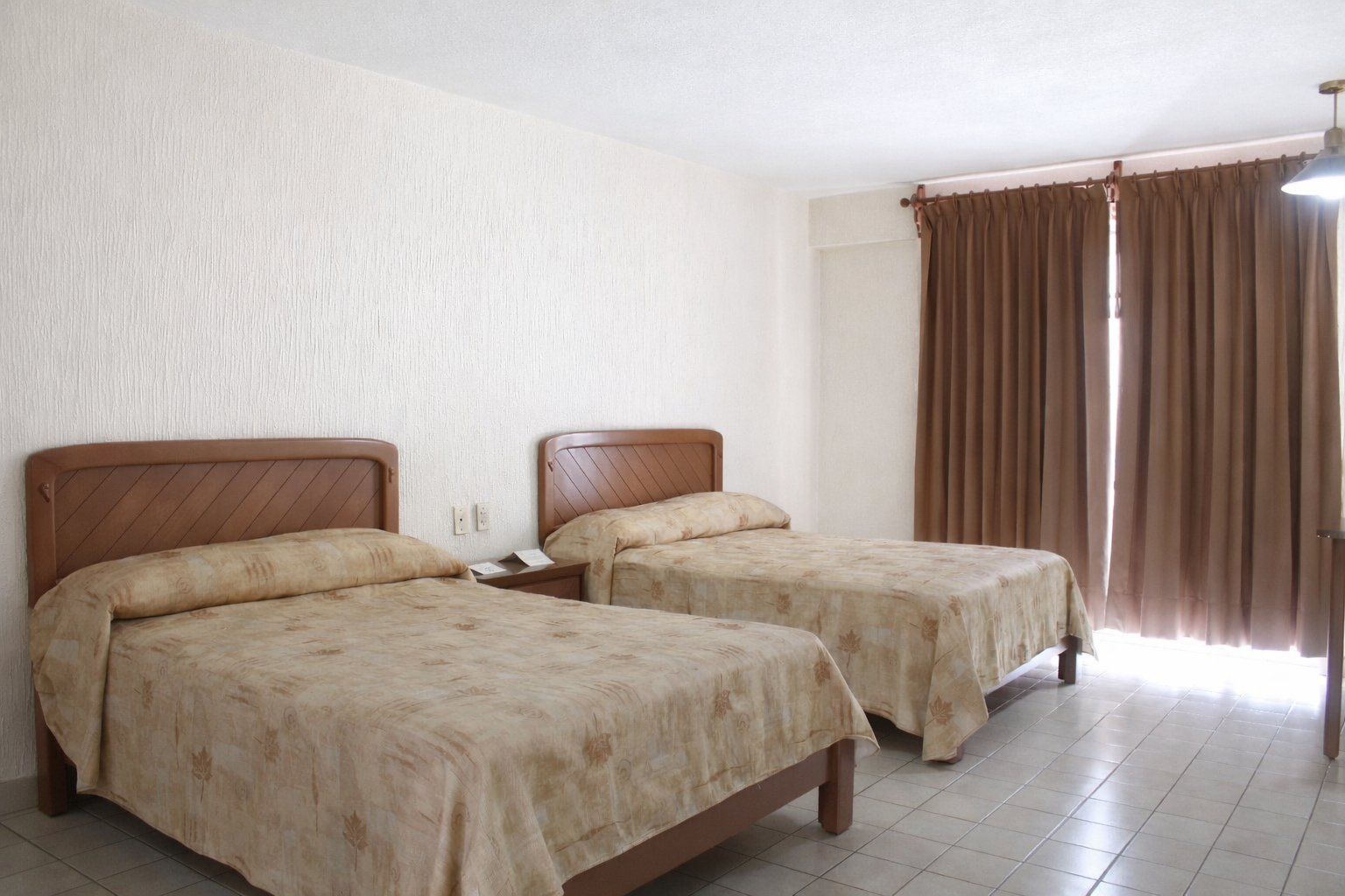 Dos camas individuales con cabeceros marrones en una habitación con cortinas marrones y paredes de color blanquecino.