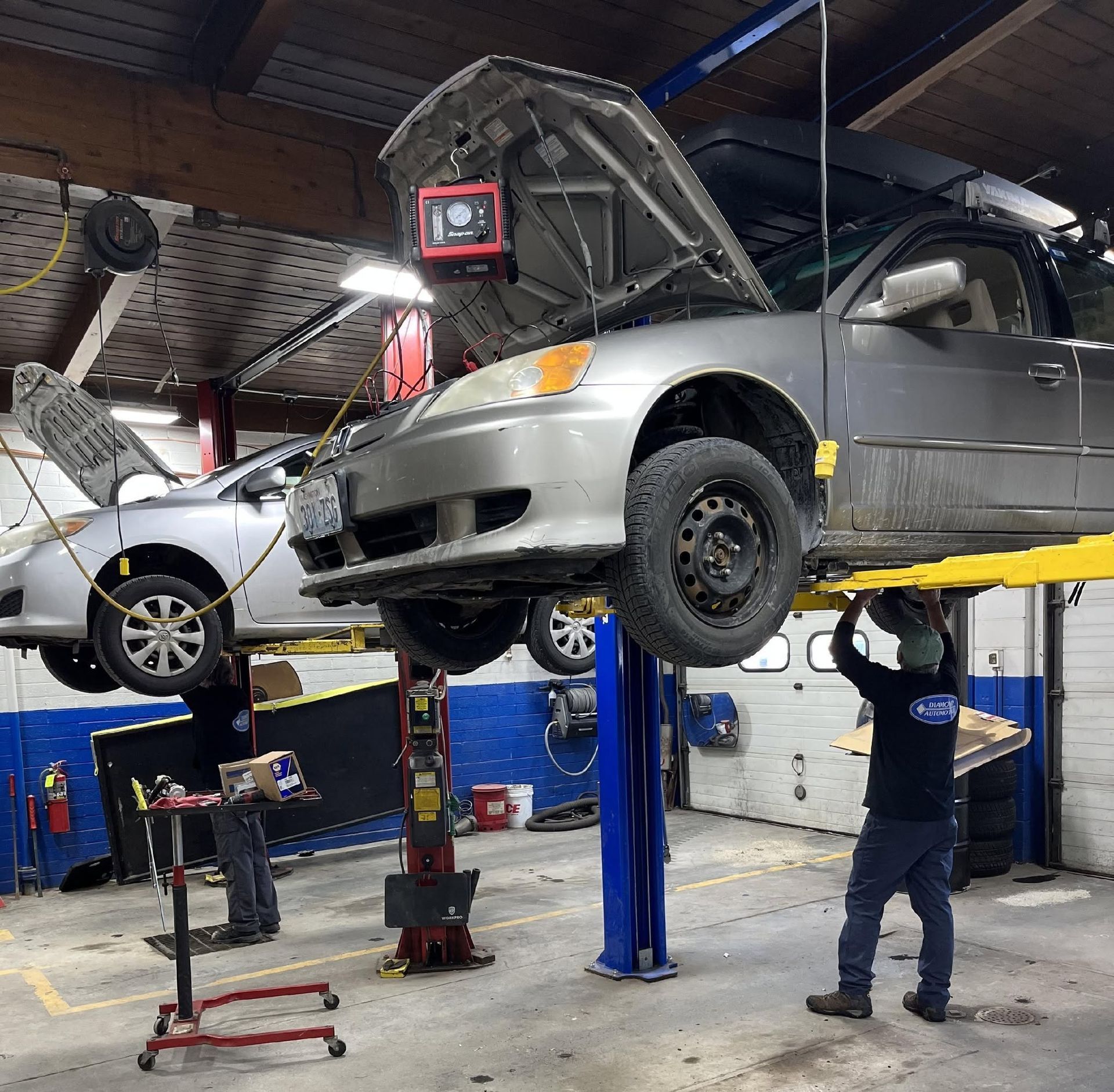 Two cars on hydraulic lifts in an auto repair garage, with a mechanic working underneath | Diamond Automotive