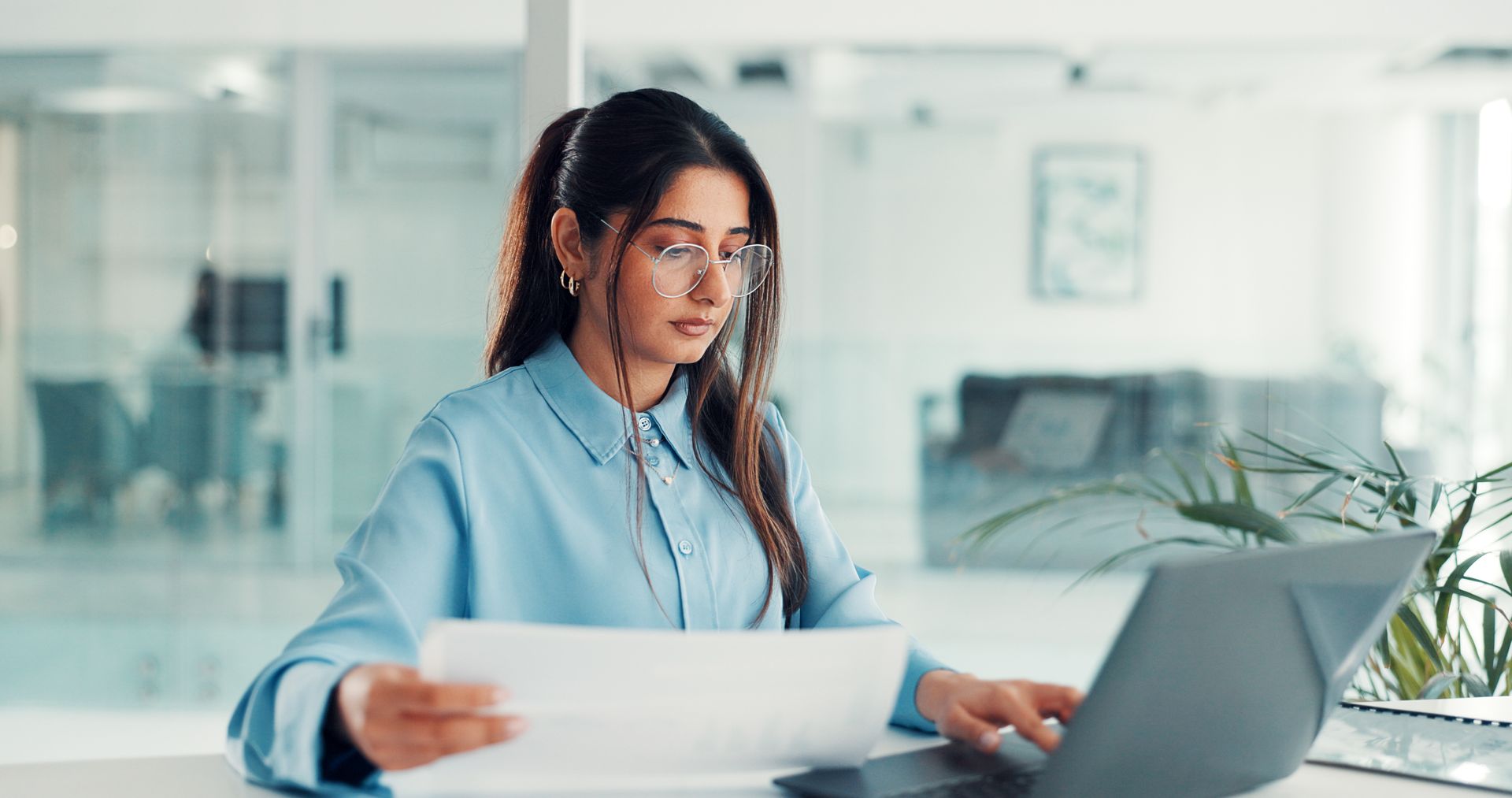 Woman in a blue shirt working on a laptop at a bright office desk, holding a document.
