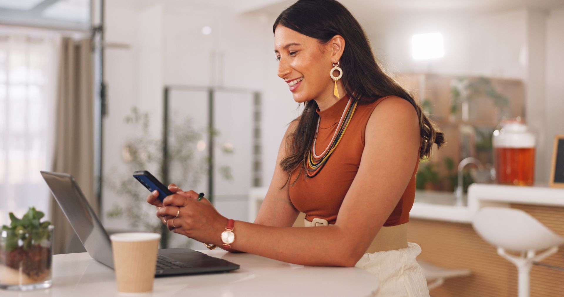 Person smiling and using a phone beside a laptop at a kitchen counter