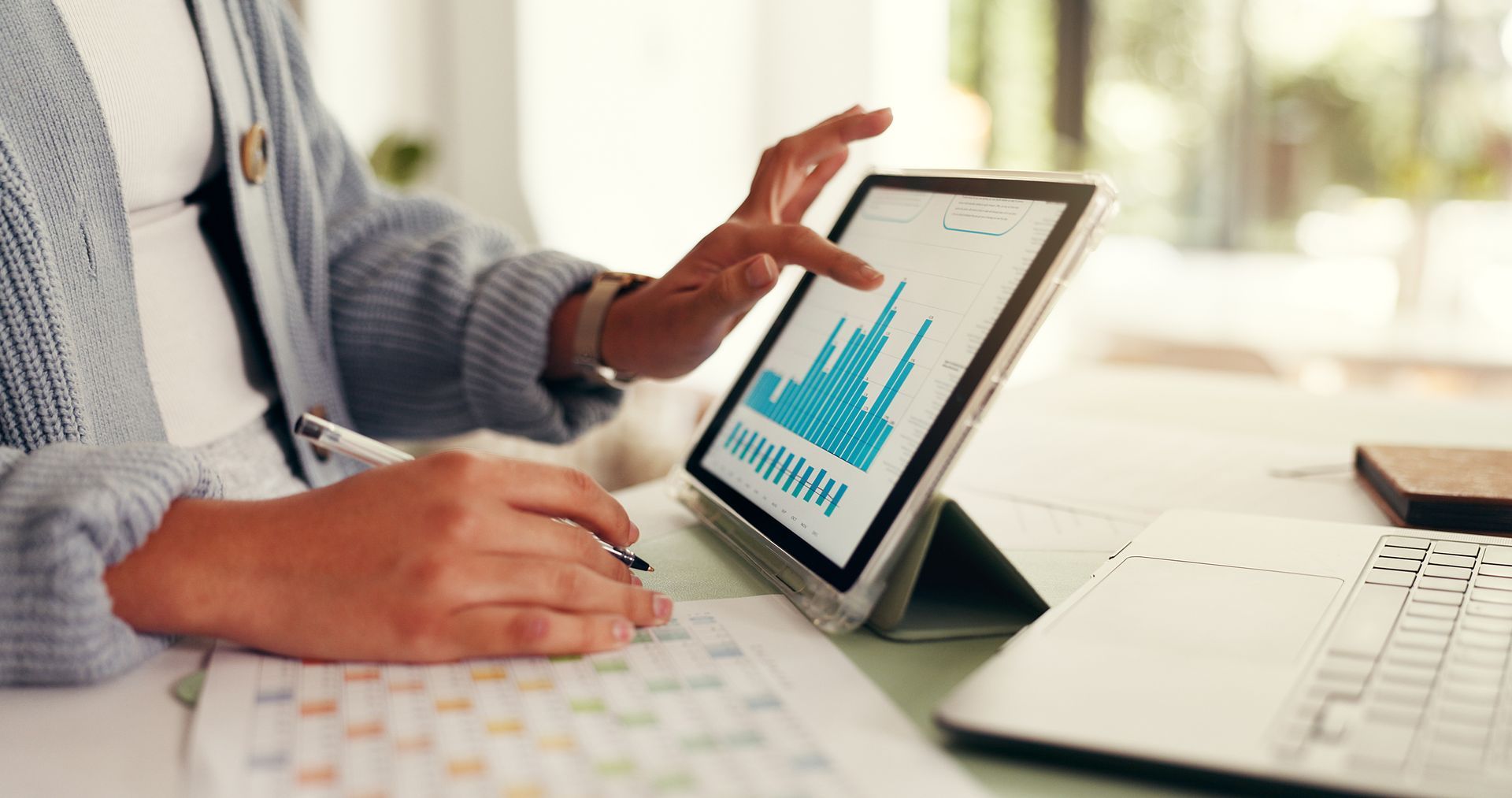 Hands using a tablet with a blue bar chart beside a laptop on a desk
