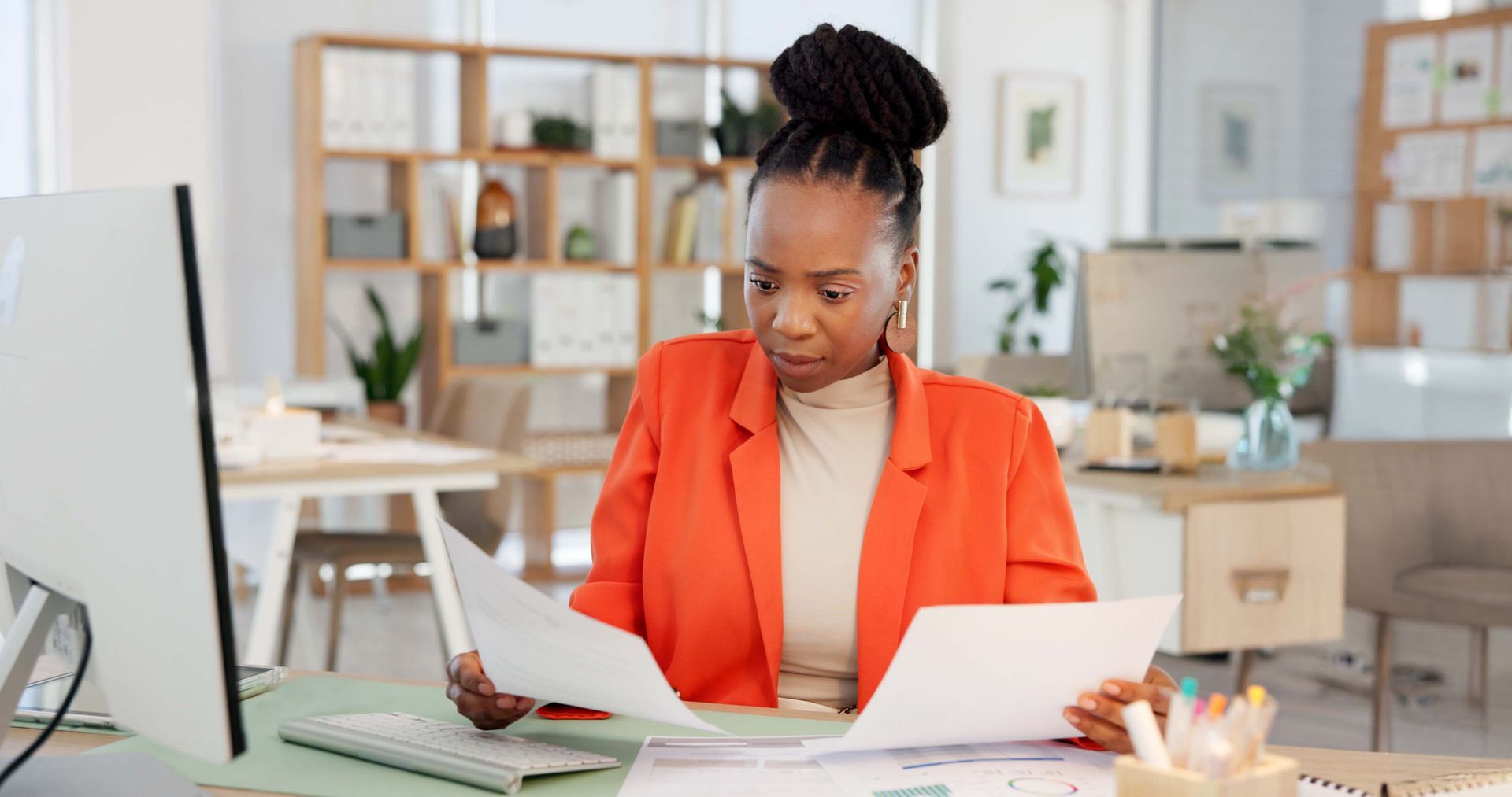 Woman in an orange blazer reviewing papers at a desk in a bright office