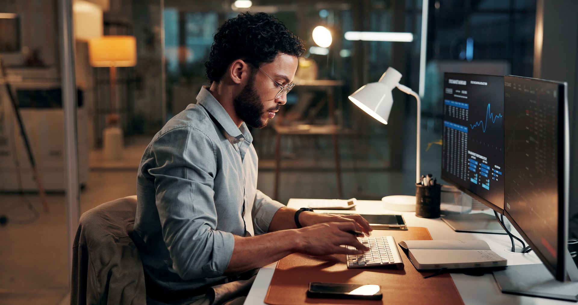 Man typing at a desk with dual monitors in a dimly lit office, focused on coding.