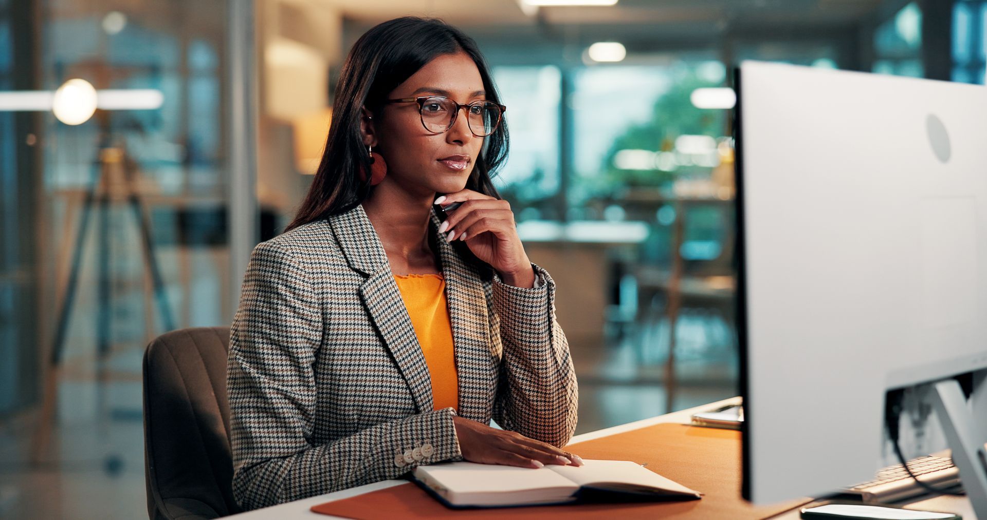 Woman in a plaid blazer sits at a desk, looking at a computer monitor in a modern office