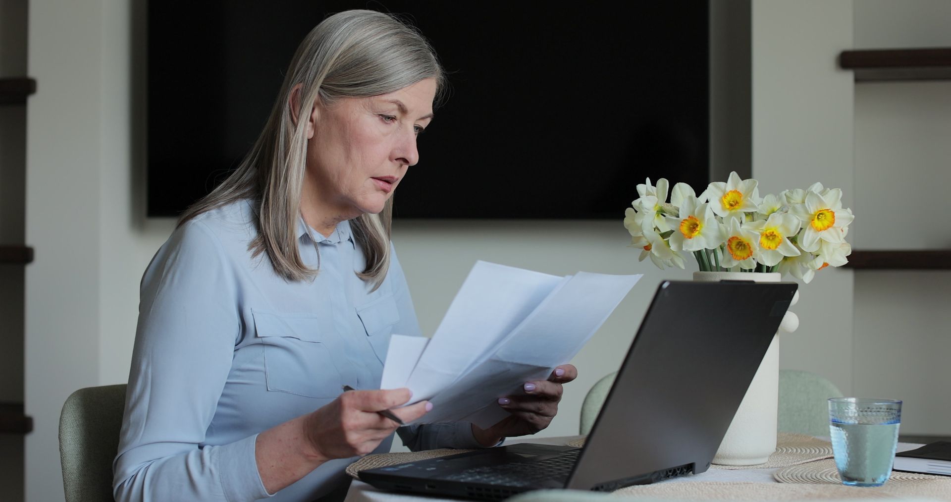 Woman reviewing papers beside a laptop on a desk with a vase of white daisies