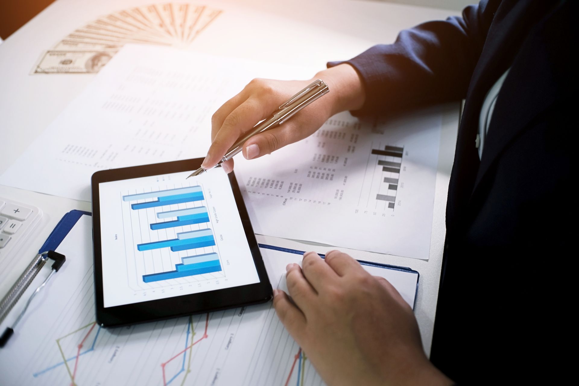Person in a suit reviews financial charts on a tablet, pointing with a pen. Money and documents are on the table.