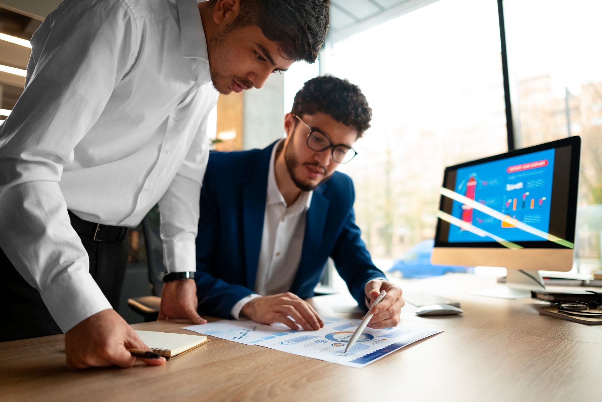 Two colleagues in professional attire collaborate over documents at a desk with a computer monitor in a bright office.