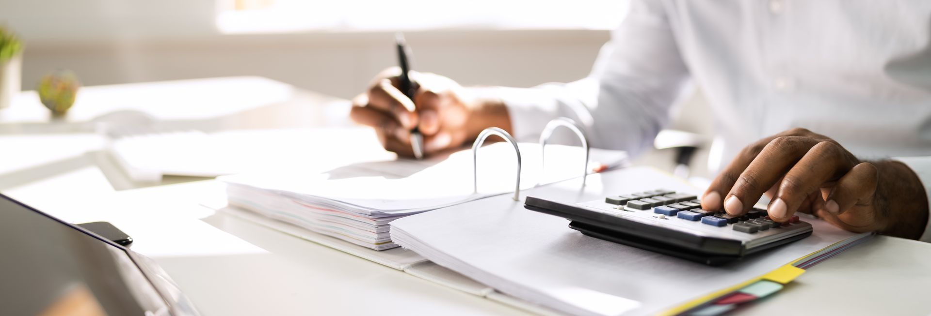 Hands working at a desk with a calculator, notebook, and pen in a bright office setting