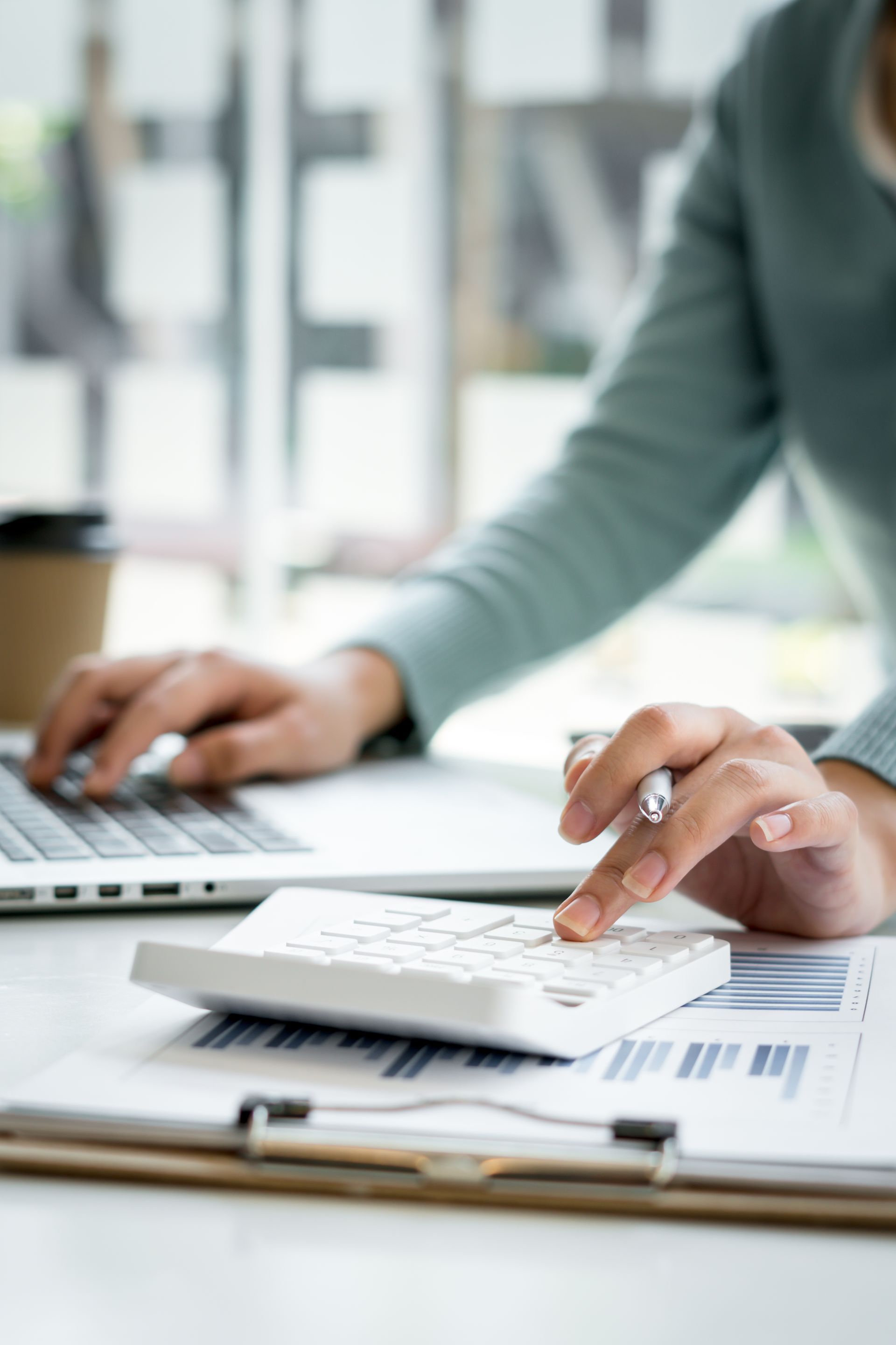 Person using a calculator and laptop, working with charts in a bright office.