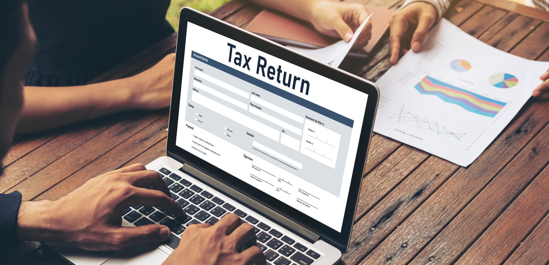 Person filling out a tax return on a laptop, with another person looking at paperwork on a wooden table.