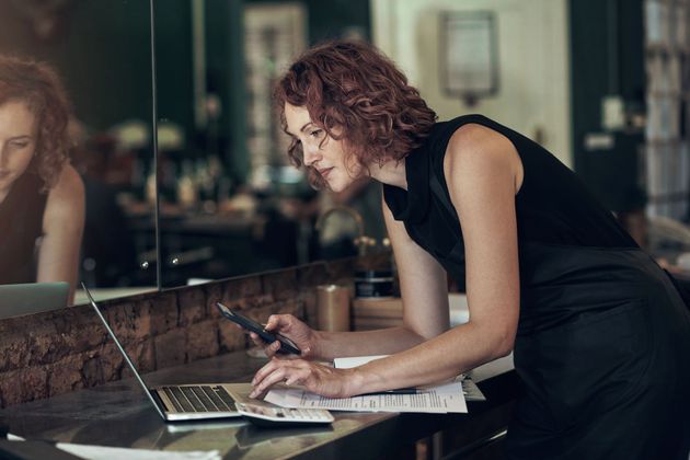 Person in a black dress leans over a laptop, writing notes at a table in a modern office